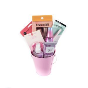 The Women's Beauty Basket - Small includes a variety of beauty products, such as nail polish, a makeup brush, Sweet Pea body spray, and other skincare items. The bucket's handle is visible, and the products are arranged neatly in their pastel-colored packaging.