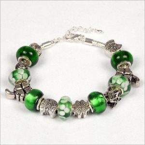 The Celtic Princess Bracelet, featuring a silver chain adorned with green and white glass beads along with various silver charms like a four-leaf clover, a butterfly, and a flower, is beautifully displayed on a white surface.