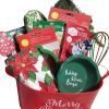 A festive red Automotive Basket labeled "Merry Christmas" holds a holiday-themed assortment, much like a cheerful basket. Inside, you'll find a poinsettia-patterned towel, cookie cutters, cupcake cups, a red and green whisk, holiday paper plates, and a green bowl with "Baking Spirits Bright" written on it.