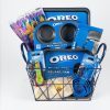 The "Everyday Baking" gift basket is filled with must-have baking essentials, including Oreo-themed items such as a 6-cup muffin pan, a square pan, a rubber spatula, and measuring spoons. Brightly colored paper cups and napkins provide a vibrant contrast to the plain white background they are presented against.