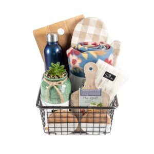 The "Kitchen & Home Medium" basket in black contains a variety of items, including a cutting board, a blue water bottle, a plaid oven mitt, a rolled-up blanket, a jar with a succulent, small kitchen tools, and tea packets. This medium-sized basket is beautifully arranged to present a rustic home kitchen gift set.