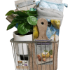 An automotive basket contains potted greenery, a cookbook, a wooden cutting board, an oven mitt, a kitchen towel, and a small wooden sign. The items are neatly arranged in the basket, making it a practical yet decorative kitchen gift set.