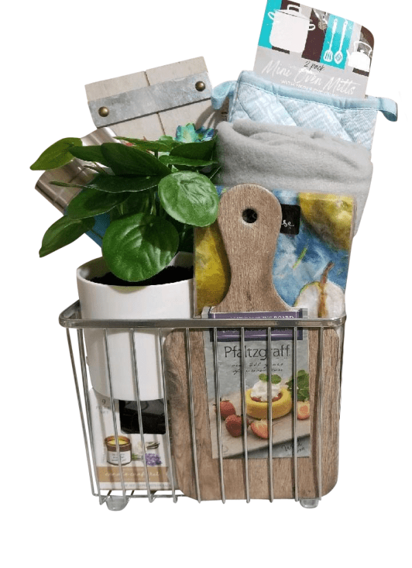 An automotive basket contains potted greenery, a cookbook, a wooden cutting board, an oven mitt, a kitchen towel, and a small wooden sign. The items are neatly arranged in the basket, making it a practical yet decorative kitchen gift set.