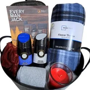 The Automotive Basket holds body washes, a gray face towel, a plaid fleece throw blanket, a red candle, and a red bath sponge. Positioned behind the basket is an Every Man Jack product brochure. The metallic basket with black trim exudes rugged elegance with its automotive charm.