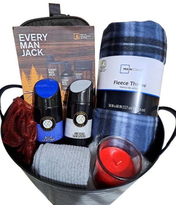 The Automotive Basket holds body washes, a gray face towel, a plaid fleece throw blanket, a red candle, and a red bath sponge. Positioned behind the basket is an Every Man Jack product brochure. The metallic basket with black trim exudes rugged elegance with its automotive charm.