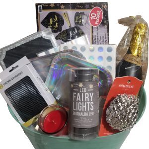 An Automotive Basket, filled with New Year's Eve celebration items, including LED fairy lights, black drink stirrers, a candle, shiny party hats, a pack of napkins, a notebook with a holographic cover, a bottle of sparkling wine, and a glittery silver ornament—the perfect way to rev up your festivities.