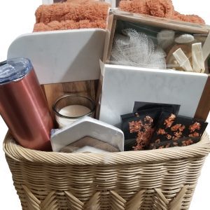 The Premium Kitchen & Home basket features a collection of exquisite items such as a copper-colored tumbler, a white candle, vibrant orange towels, luxurious skincare products, a soft white loofah, and delectable brown snacks. Some items are thoughtfully wrapped in clear or white packaging. Ideal for adding an elegant touch to your home decor. The background is set against a crisp white backdrop.