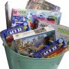 A large light green Automotive Basket filled with various jigsaw puzzles and two boxes of Act II microwave popcorn (butter flavor). The puzzles feature different themes, including birds, a snowy village, and abstract designs, with pieces ranging from 750 to 1000.