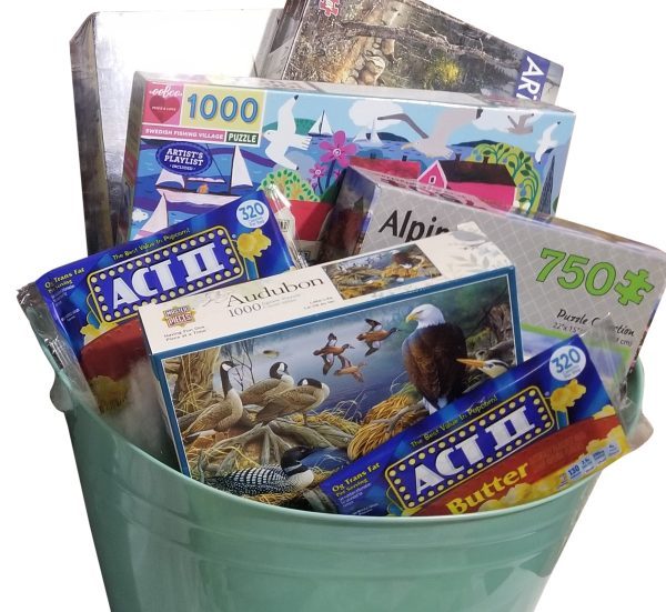 A large light green Automotive Basket filled with various jigsaw puzzles and two boxes of Act II microwave popcorn (butter flavor). The puzzles feature different themes, including birds, a snowy village, and abstract designs, with pieces ranging from 750 to 1000.