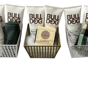 Three metal baskets containing Bulldog Original Skincare for Men products and various cloth items. The left and right baskets are white, while the center basket is gold. This arrangement complements an Epic Women's Spa Basket collection perfectly, with predominantly white packaging featuring brown lettering.