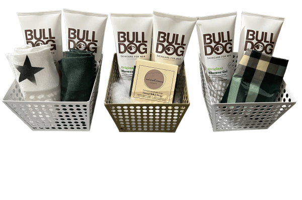 Three metal baskets containing Bulldog Original Skincare for Men products and various cloth items. The left and right baskets are white, while the center basket is gold. This arrangement complements an Epic Women's Spa Basket collection perfectly, with predominantly white packaging featuring brown lettering.