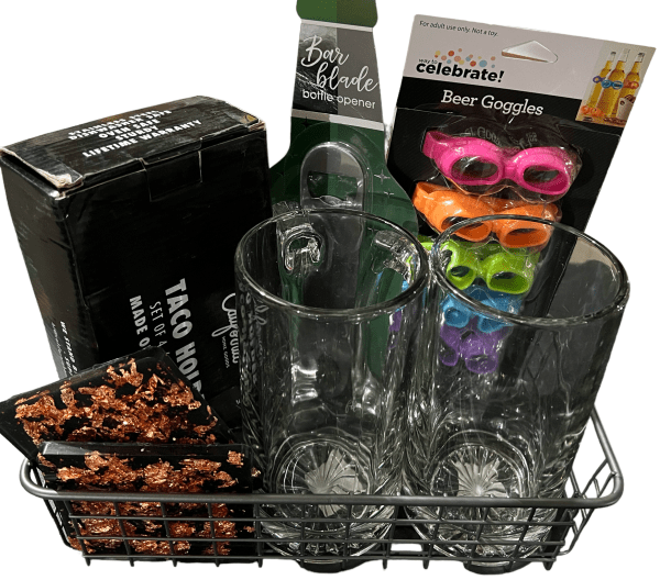 The Epic Women's Spa Basket includes two tall glasses, four colorful beer goggles, a black Taco Holders set box, a Bar Blade bottle opener, and a decorative item with a black base and metallic rose gold flakes. All items are beautifully arranged in a chic metal wire basket.