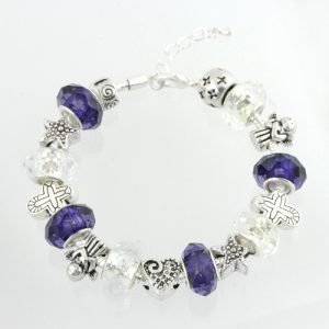 The Victory - He is Risen Bracelet, a silver charm bracelet adorned with purple, white, and clear beads interspersed with various charms including stars, crosses, and hearts, is highlighted in Christian jewelry collections. It is displayed in a circular shape on a white background to accentuate its intricate design.