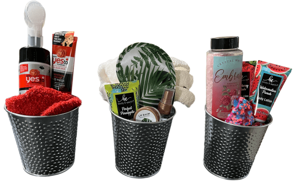 Three gift baskets with self-care products in black perforated metal containers. The left basket, labeled as the Epic Women's Spa Basket, contains a red towel, face mask, and brush. The middle basket includes a tropical-themed dish, lip balm, and scrunchie. The right basket offers lotion, bath salt, and watermelon-themed items.