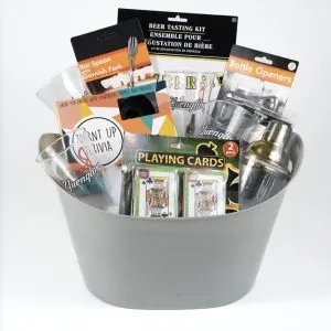 The Adult Game Night Basket features a gray bucket filled with an array of bar-themed items, including a beer tasting kit, a bar spoon and garnish fork, bottle openers, playing cards, drinking glasses, and a cocktail shaker—all set against a white background. It's the perfect addition to any adult game night.