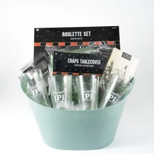 The Adult Gambling Night Basket features a green bucket filled with assorted items, including two tall beer glasses labeled "Yuchip IPA," a box labeled "Roulette Set," a craps table cover, and other small accessories—perfect for an adult night of gambling fun. The background is white.