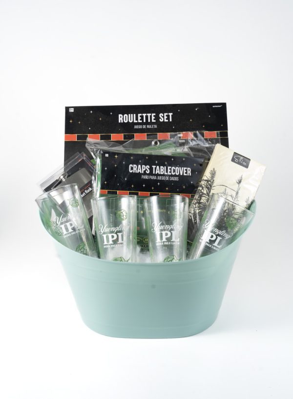 The Adult Gambling Night Basket features a green bucket filled with assorted items, including two tall beer glasses labeled "Yuchip IPA," a box labeled "Roulette Set," a craps table cover, and other small accessories—perfect for an adult night of gambling fun. The background is white.