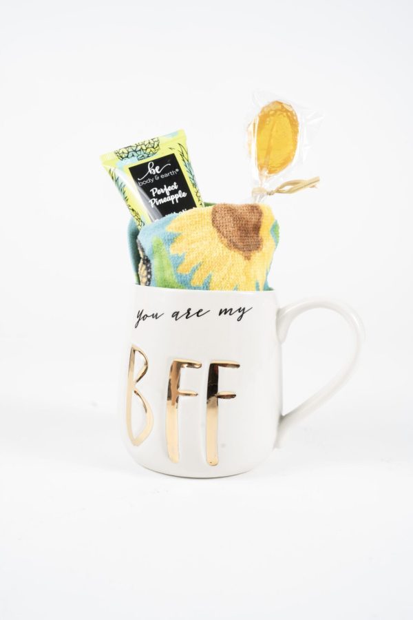 The You are my BFF Mug Gift Basket with Free Food Certificate features a white mug adorned with gold letters spelling "BFF" and the text "you are my" at the top, filled with a yellow lollipop, a pineapple-flavored candy packet, and a colorful sunflower-themed towel. This delightful gift set is the perfect addition to any gift basket and stands out beautifully against a plain white background.