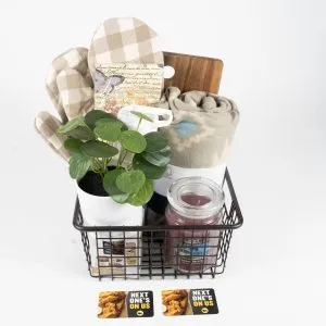 A Black Wire Basket filled with a small potted plant, a beige checkered oven mitt, a wooden cutting board, a rolled-up blanket, and a jar candle. Two "Next One's On Us" gift cards featuring images of food are placed in front of the basket, making it the perfect BW3's Kitchen & Home Basket - Includes $50 in BW3's Gift Cards.