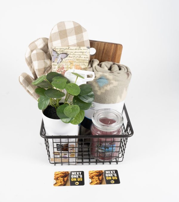 A Black Wire Basket filled with a small potted plant, a beige checkered oven mitt, a wooden cutting board, a rolled-up blanket, and a jar candle. Two "Next One's On Us" gift cards featuring images of food are placed in front of the basket, making it the perfect BW3's Kitchen & Home Basket - Includes $50 in BW3's Gift Cards.