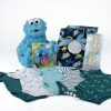 Displayed is the Adult Gambling Night Basket, which includes a blue Cookie Monster plush toy** for luck, baby clothes in shades of teal and white with various patterns **for cozy vibes**, a matching blanket **for comfort**, a pack of baby bath time essentials **to unwind after the game**, and a blue and green teething toy—all neatly arranged in this charming basket.