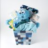 The Adult Gambling Night Basket contains a blue plush toy resembling a monster with googly eyes, a rolled-up blanket with animal prints, a set of baby pacifiers, baby bodysuits, a pair of socks, and a teething toy, all arranged in a blue and gray patterned container.