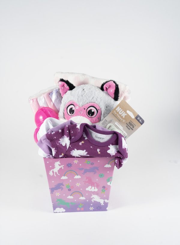 An "Adult Gambling Night Basket" features a purple gift box adorned with unicorns and rainbows. Inside, it contains a plush animal with large pink eyes, a purple and white baby outfit, a set of pink baby utensils, and a NUK pacifier in its packaging. Under the night sky, the items are arranged neatly in the box.