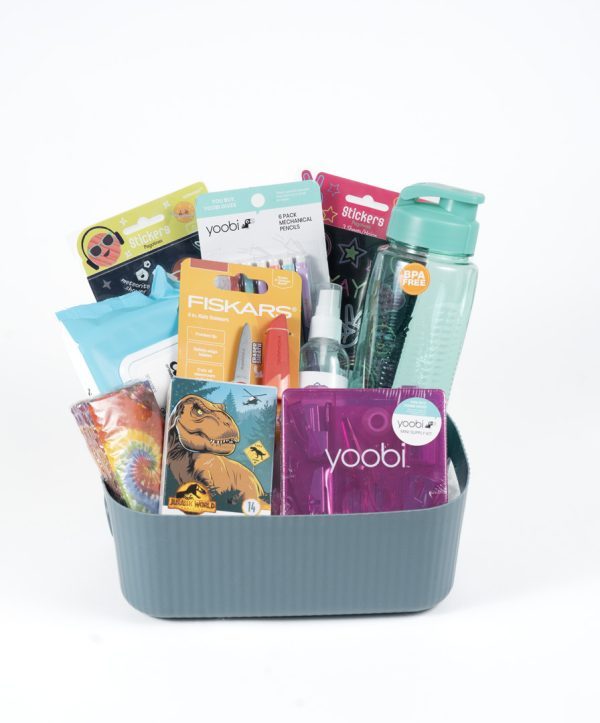 A gray basket labeled "Adult Gambling Night Basket" sits against a white background, containing various themed supplies including a water bottle, pencils, markers, stickers, scissors, glue stick, a pack of moist wipes, a small dinosaur-covered notebook, and colorful notepads. It's perfect for organizing your child's essentials.