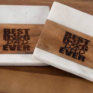 Placed on a wooden surface, the Pair of Best Dog Dad Ever Square Coasters feature a combination of wood and white marble. These square coasters are engraved with "Best Dog Dad Ever" and have a charming paw print between "Dog" and "Dad," exuding a simple, rustic charm.