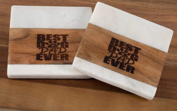 Placed on a wooden surface, the Pair of Best Dog Dad Ever Square Coasters feature a combination of wood and white marble. These square coasters are engraved with "Best Dog Dad Ever" and have a charming paw print between "Dog" and "Dad," exuding a simple, rustic charm.