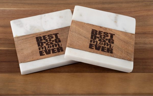 A Pair of Best Dog Mom Ever Square Coasters, featuring a design of light wood and white marble with each coaster engraved with the phrase "Best Dog Mom Ever" and a paw print replacing the "O" in "Dog," placed on a wooden surface.