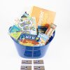 A blue gift basket, known as the "Big Air Trampoline & Pizza Entertainment Basket," is filled with assorted items including snacks, a book titled "Haggin' Words," a game called "Dream On!", popcorn, candy, and various entertainment coupons. Three pizza coupons are displayed in front of the basket.