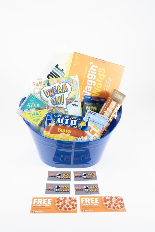 A blue gift basket, known as the "Big Air Trampoline & Pizza Entertainment Basket," is filled with assorted items including snacks, a book titled "Haggin' Words," a game called "Dream On!", popcorn, candy, and various entertainment coupons. Three pizza coupons are displayed in front of the basket.