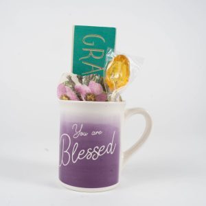 The "You are Blessed Mug Gift Basket with Free Food Certificate" features a stunning white and purple gradient Blessed Mug inscribed with "You are Blessed." Inside, you'll find a small green plaque reading "Grateful," along with a pink and purple plush toy and a honey lollipop wrapped in plastic. This delightful ensemble is showcased against a plain background, creating an exquisite gift basket.