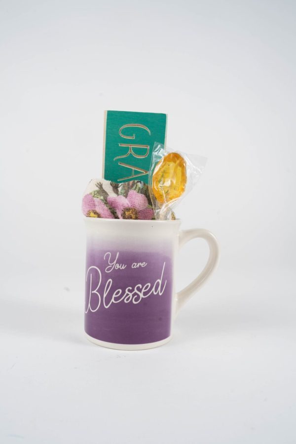 The "You are Blessed Mug Gift Basket with Free Food Certificate" features a stunning white and purple gradient Blessed Mug inscribed with "You are Blessed." Inside, you'll find a small green plaque reading "Grateful," along with a pink and purple plush toy and a honey lollipop wrapped in plastic. This delightful ensemble is showcased against a plain background, creating an exquisite gift basket.