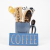 A blue ceramic Spooner Mug from the Spooner Mug Gift Basket with Free Food Certificate holds a packaged cookie and a brown spoon. In front of the mug is a blue rectangular sign with the word "COFFEE" in white capital letters. The background is plain white.