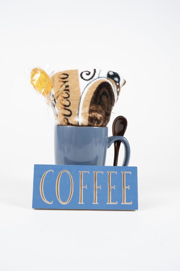 A blue ceramic Spooner Mug from the Spooner Mug Gift Basket with Free Food Certificate holds a packaged cookie and a brown spoon. In front of the mug is a blue rectangular sign with the word "COFFEE" in white capital letters. The background is plain white.