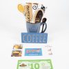 A blue Spooner Mug from the "Spooner Mug Gift Basket with Free Food Certificate" collection, filled with a rolled-up coffee-themed towel, is placed next to a wooden sign that says "COFFEE." Below are a Wendy's gift card, a pack of McDonald's hazelnut iced coffee sachets, and two $10 Tradewinds coupons, making it the perfect gift basket for any coffee lover.