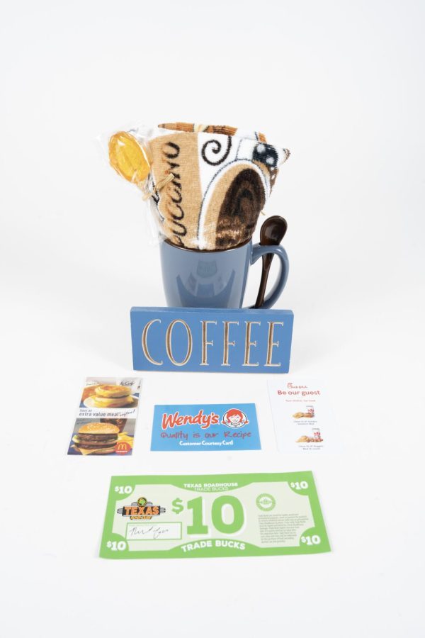 A blue Spooner Mug from the "Spooner Mug Gift Basket with Free Food Certificate" collection, filled with a rolled-up coffee-themed towel, is placed next to a wooden sign that says "COFFEE." Below are a Wendy's gift card, a pack of McDonald's hazelnut iced coffee sachets, and two $10 Tradewinds coupons, making it the perfect gift basket for any coffee lover.