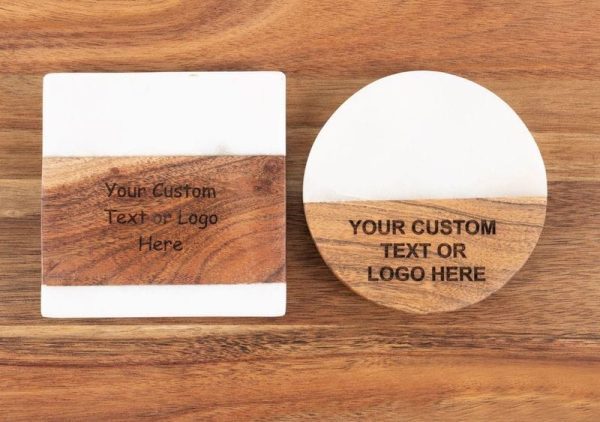 The Personalized Kitchen & Home Gift Basket includes distinguished square and round custom coasters with a wooden and white split design—the square coaster is paired with a round one. Both coasters elegantly feature the text "Your Custom Text or Logo Here" engraved on the wooden portion. They rest gracefully on a sophisticated wooden cutting board, complementing the basket's charm.