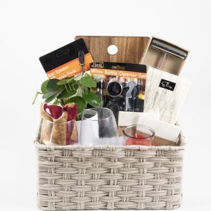 The Personalized Celebration Basket with 2 Custom Wine Glasses & Cutting Board features a wicker basket filled with a bar spoon/stirrer set, a wine bottle opener set, a small potted plant, two custom wine glasses, a red candle, a small wooden cutting board, and a box of snacks. These items are neatly arranged within the basket.