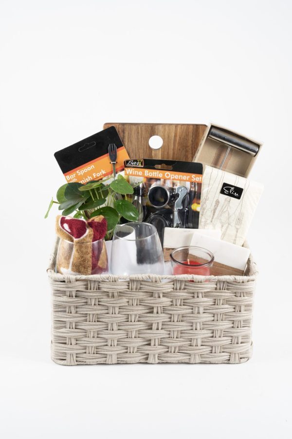 The Personalized Celebration Basket with 2 Custom Wine Glasses & Cutting Board features a wicker basket filled with a bar spoon/stirrer set, a wine bottle opener set, a small potted plant, two custom wine glasses, a red candle, a small wooden cutting board, and a box of snacks. These items are neatly arranged within the basket.