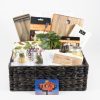 A woven basket is filled to the brim with various kitchen essentials—a wooden cutting board, a bar spoon with a garnish fork, a bottle opener set, a potted plant, and a tea towel adorned with a grape pattern. It also includes $75 in Texas Roadhouse Certificates and other Home & Wine items for the perfect Celebration Basket.