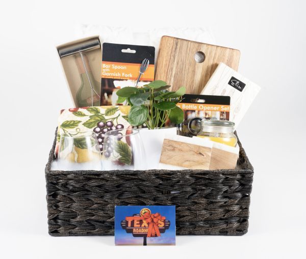 A woven basket is filled to the brim with various kitchen essentials—a wooden cutting board, a bar spoon with a garnish fork, a bottle opener set, a potted plant, and a tea towel adorned with a grape pattern. It also includes $75 in Texas Roadhouse Certificates and other Home & Wine items for the perfect Celebration Basket.