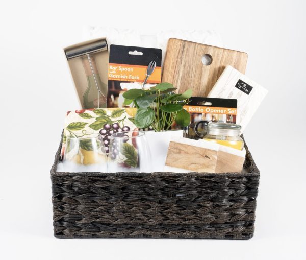 A Personalized Kitchen & Home Gift Basket, featuring a black woven design, is filled with an array of kitchen and bar essentials. Inside, you'll find a bottle opener set, a custom cutting board, a potted plant, custom glasses adorned with fruit designs, a bar spoon with a garnish fork, and an assortment of kitchen towels. This makes for the perfect personalized kitchen gift.