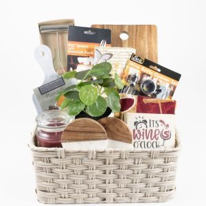 A Personalized Celebration Basket with 2 Custom Coasters & Cutting Board, filled with various items including a small potted plant, a wine bottle opener set, a bar spoon and garnish fork set, a jar candle, a cheese knife, and a red cloth napkin with custom coasters featuring the text "It's Wine O'Clock," perfect for any special occasion.