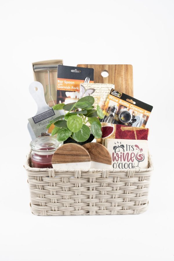 A Personalized Celebration Basket with 2 Custom Coasters & Cutting Board, filled with various items including a small potted plant, a wine bottle opener set, a bar spoon and garnish fork set, a jar candle, a cheese knife, and a red cloth napkin with custom coasters featuring the text "It's Wine O'Clock," perfect for any special occasion.