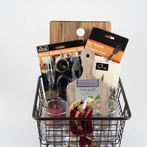 The Personalized Celebration Basket Medium with Custom Cutting Board features a black wire basket filled with various kitchen essentials, including a wine bottle opener set, a bar spoon and garnish fork, a small glass candle, two shot glasses, and a custom cutting board. Also included is a cheese board adorned with red and green peppers and finished with a ribbon for that extra touch of personalization.