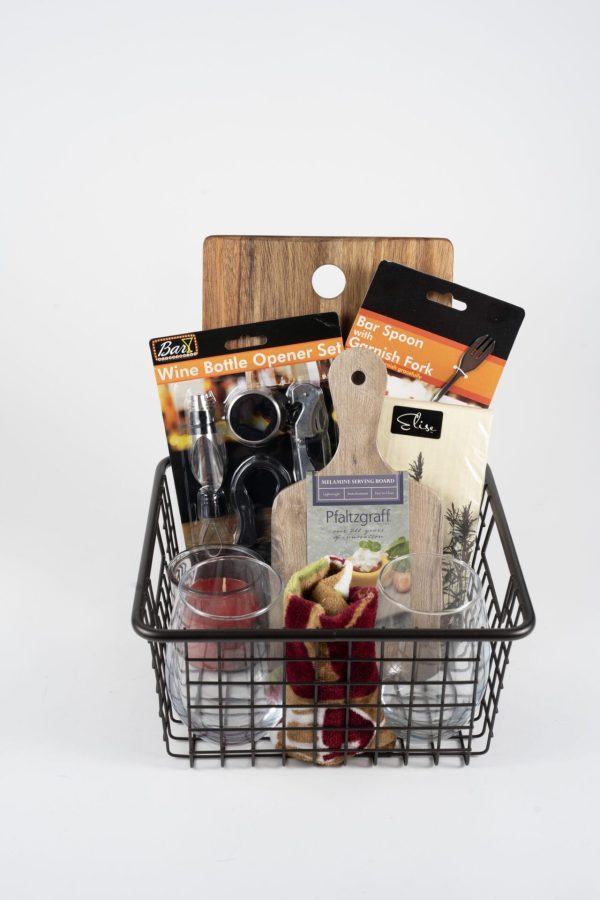 The Personalized Celebration Basket Medium with Custom Cutting Board features a black wire basket filled with various kitchen essentials, including a wine bottle opener set, a bar spoon and garnish fork, a small glass candle, two shot glasses, and a custom cutting board. Also included is a cheese board adorned with red and green peppers and finished with a ribbon for that extra touch of personalization.