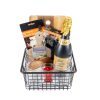 The Personalized Celebration Basket Medium with 2 Custom Coasters includes a metal wire basket that holds a bottle of sparkling wine, a wooden cutting board with a visible handle, a set of bar spoon and garnish fork, a wine bottle opener, a small wooden jar, two glass candles, and custom coasters. Stylish packaging labels are also part of the presentation.